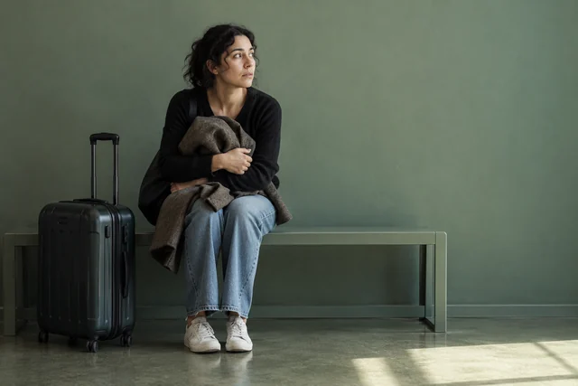 Una mujer adulta recién llegada está sentada junto a una maleta cerrada, abraza su abrigo y mira alrededor buscando una referencia; un banco sencillo y mucho espacio vacío.