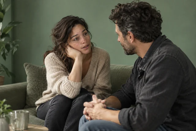 Una mujer adulta en una reunión social mantiene una conversación cercana con otro adulto; su expresión mezcla necesidad de conexión y vulnerabilidad. Ambos completamente vestidos, composición no sexualizada.