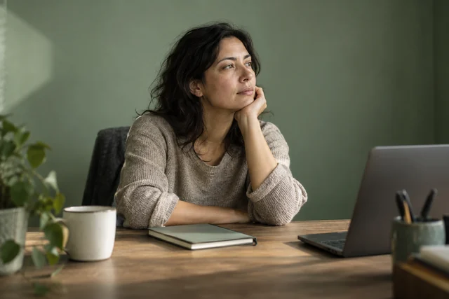 Una mujer adulta sentada frente a un escritorio deja de mirar su trabajo y observa fuera de cuadro, soñadora y distante, con un cuaderno cerrado y luz lateral.